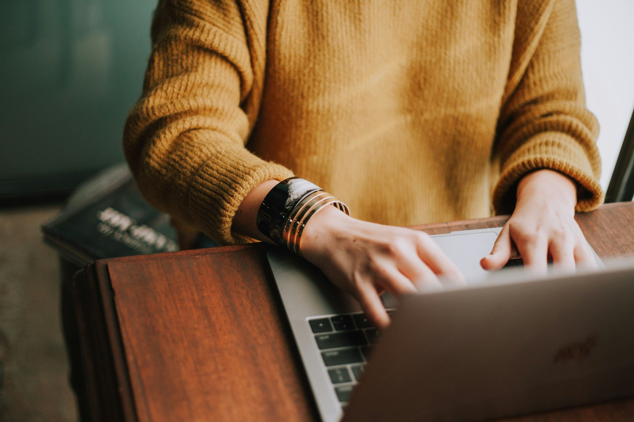 A person in a mustard sweater works on a laptop at a wooden desk